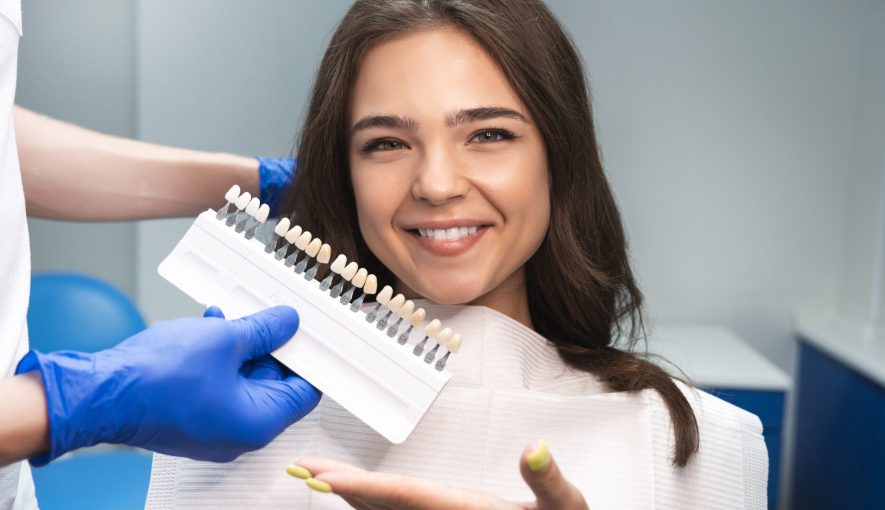 smiling brunette woman patient having appiontment in dental clinic picking up shade using tooth enamel scale held by dentist in blue gloves.